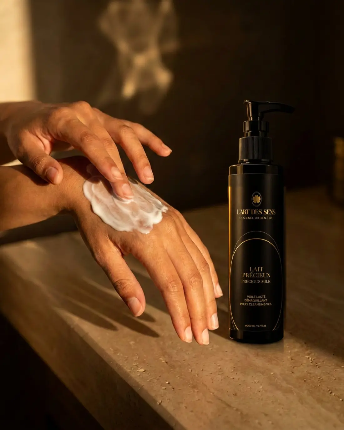 Le Lait Précieux Démaquillant L’Art des Sens — texture lactée appliquée sur le dos de la main, flacon posé sur travertin