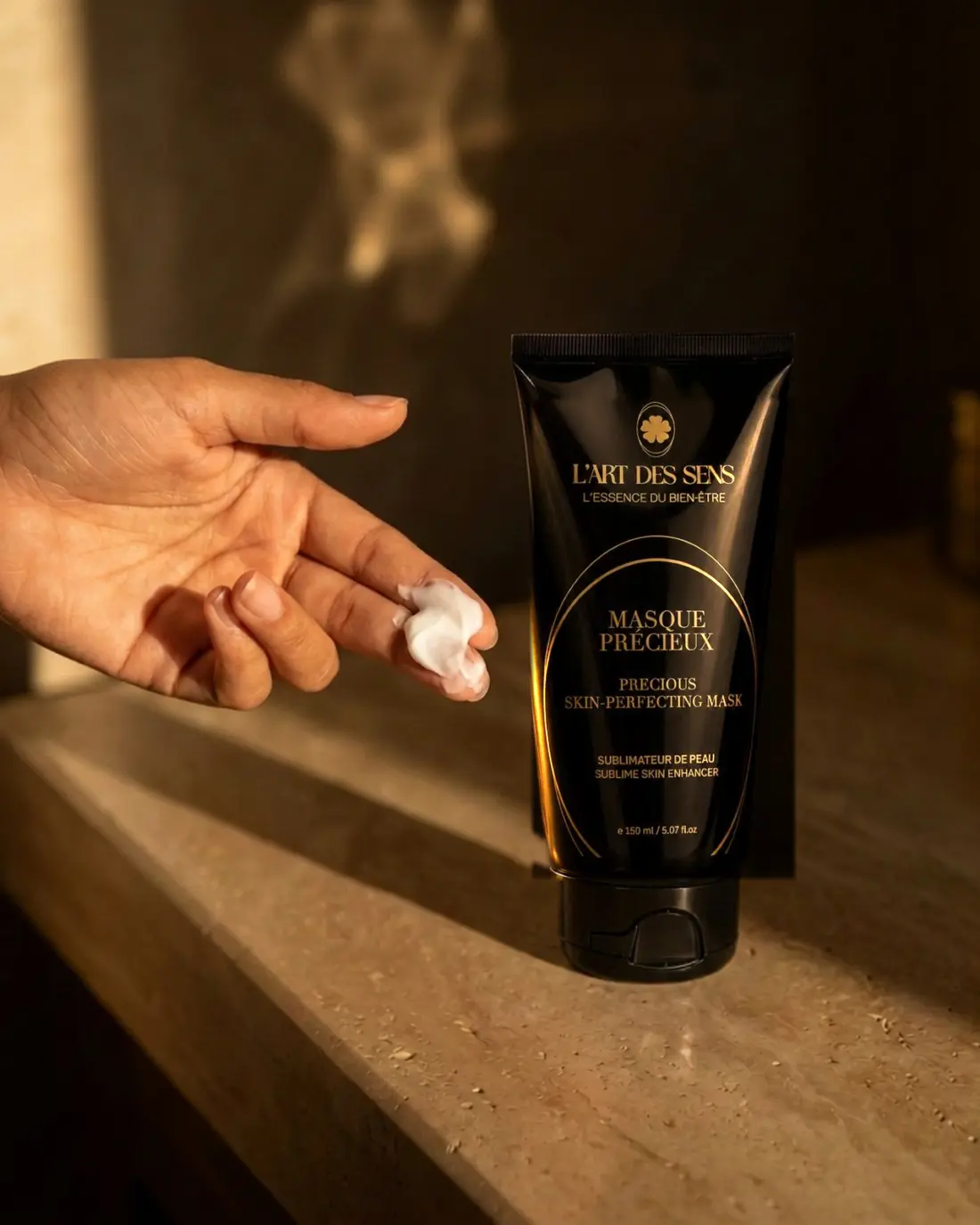 Le Masque Précieux L’Art des Sens — noisette de masque crémeux sur les doigts, tube posé sur travertin